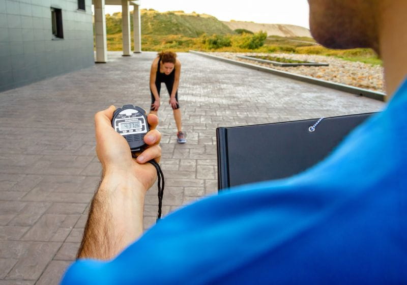 homem com um cronometro na mão e uma mulher com as mãos nos joelhos cansada de tanto correr 