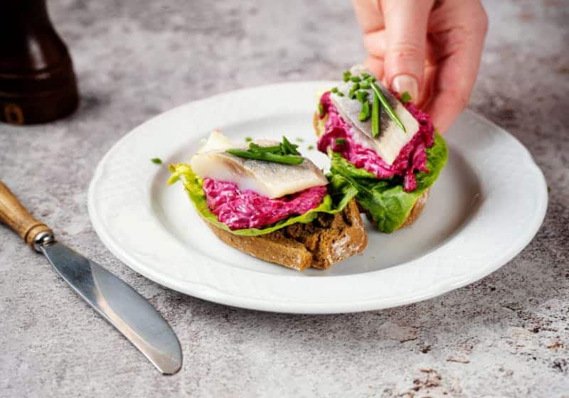 mão mexendo em um pão com coisas saudável em cima com um prato branco e uma faca ao lado 