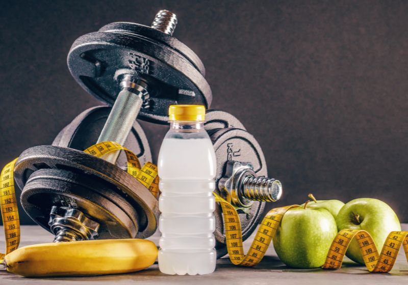 Imagem com aparelhos de musculação, frutas e uma garrafa de água