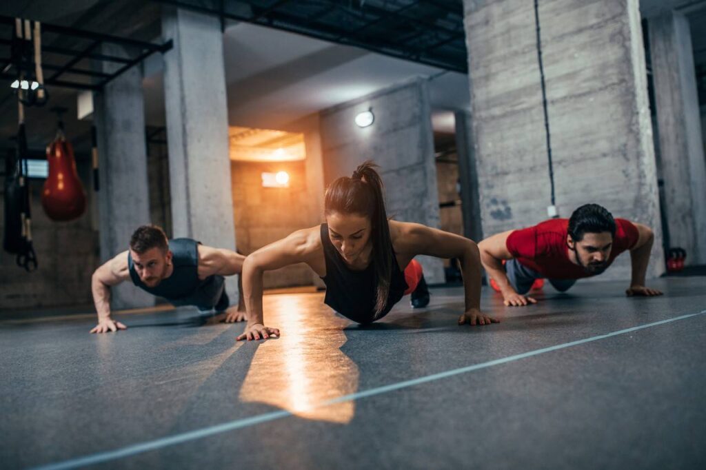 três pessoas realizando o exercício de flexão em ambiente interno de academia