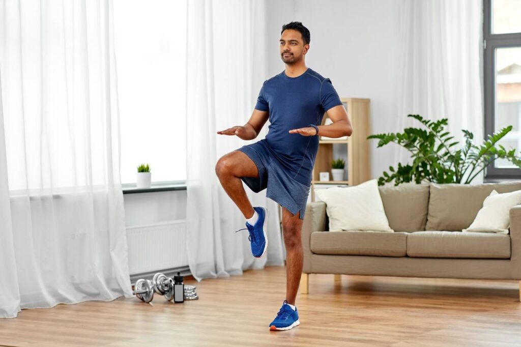 Homem fazendo exercício cardio dentro de casa, sala com sofá no tom bege, almofadas brancas, vaso de plantas e alguns pesos no chão