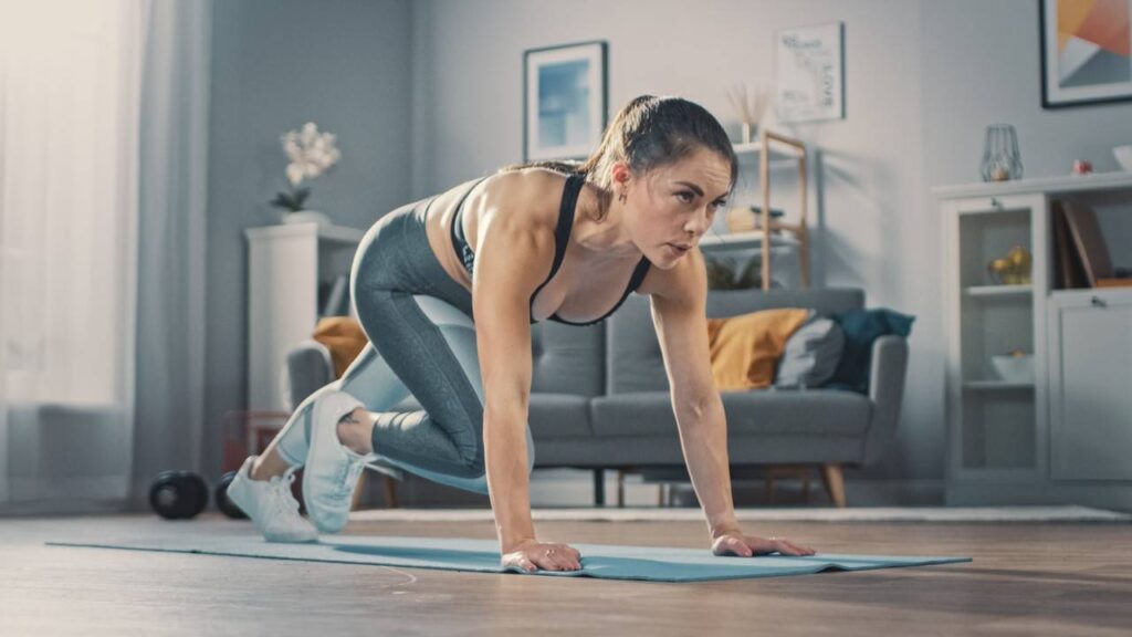 Mulher fazendo exercício cardio dentro de casa, sala com sofá azul e almofadas