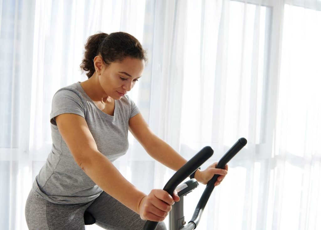 Mulher vestida com roupas de academia no tom cinza, fazendo exercício de pedal em bicicleta ergométrica.