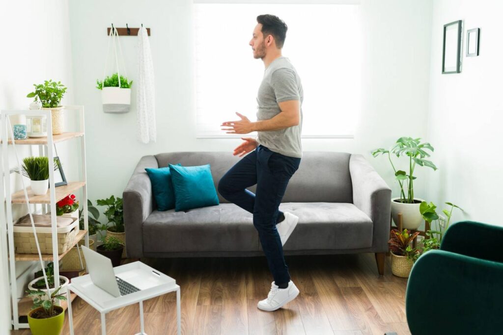 Homem fazendo exercício em sala de Tv, ao fundo sofá no tom cinza, almofadas na cor azul, vaso de plantas e quadros fixos na parede