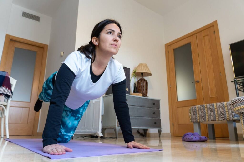 Mulher realizando atividade física em casa, com braços e uma perna apoiando ao chão, outra perna levantada