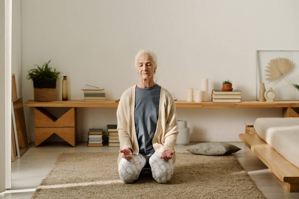 Idosa sentada ao chão com olhos fechando e praticando meditação em yoga