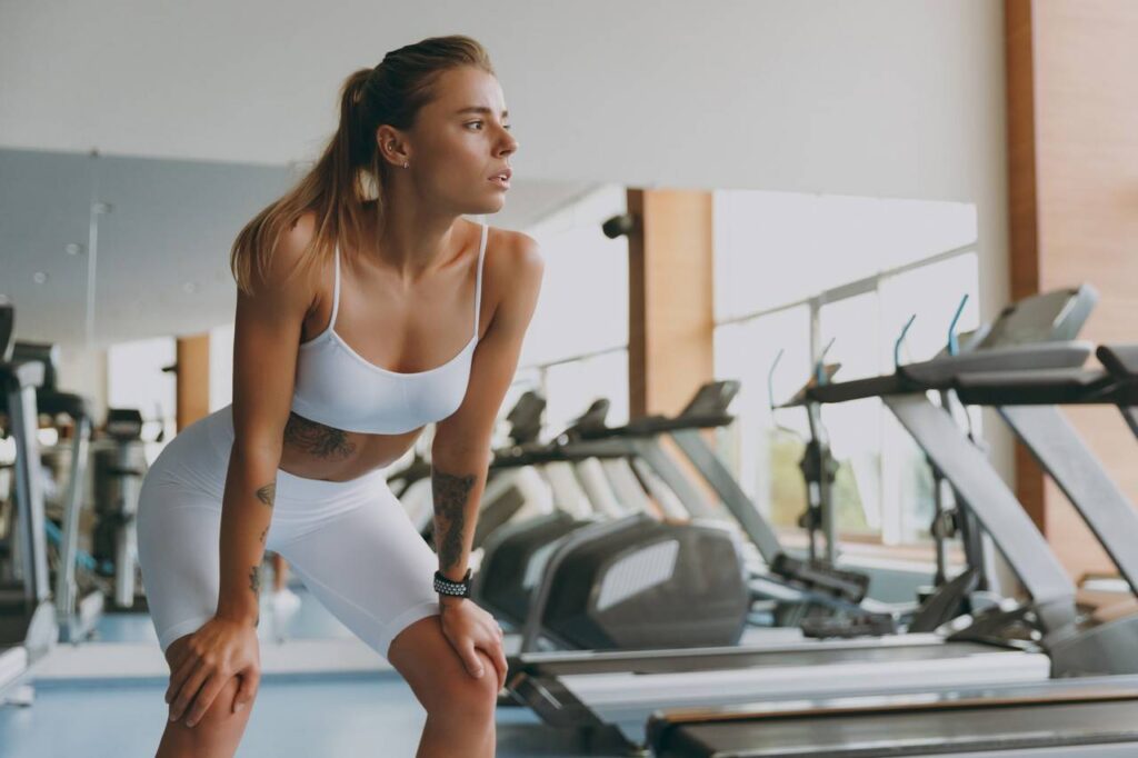 Mulher com mãos sobre os joelhos, ganhando fôlego, ao fundo várias esteiras em academia