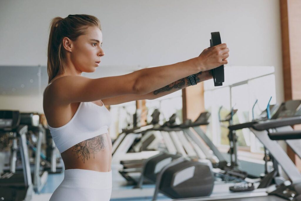 Mulher em academia segurando anilha com ambos os braços esticados