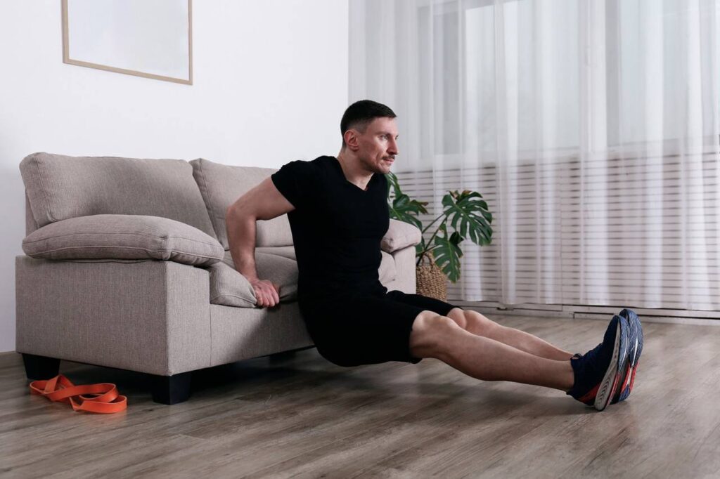Homem praticando exercício em casa, com braços apoiados no sofá e pernas esticadas, realizando triceps banco